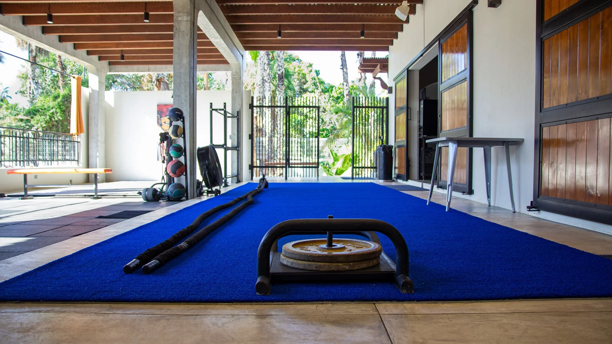 Indoor gym area with kettlebells, medicine balls, a bench, and battle ropes on a blue mat, with open doors leading outside to greenery.