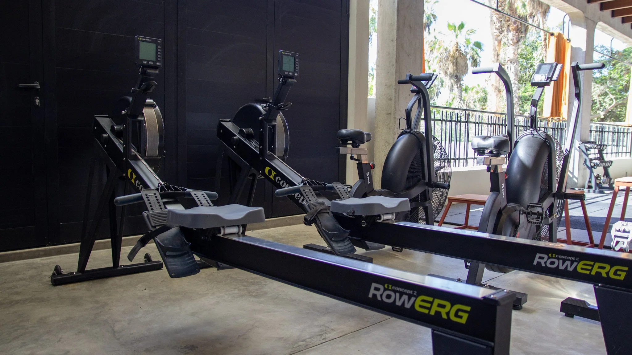 Indoor gym with rowing machines and exercise bikes facing large windows revealing palm trees outside.