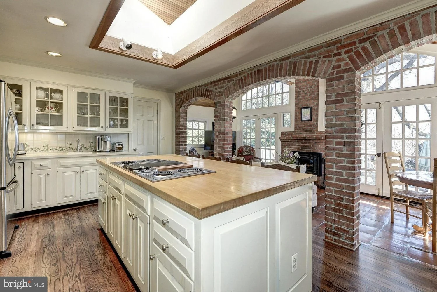 A spacious kitchen with white cabinets, a central wooden island with a cooktop, and brick arches dividing the kitchen from the dining and living area. Large windows and a skylight illuminate the space.