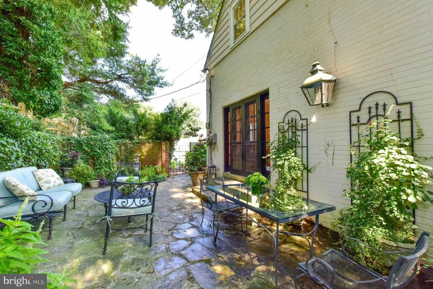 Outdoor patio with iron furniture, potted plants, and lush greenery beside a white brick house with outdoor lighting