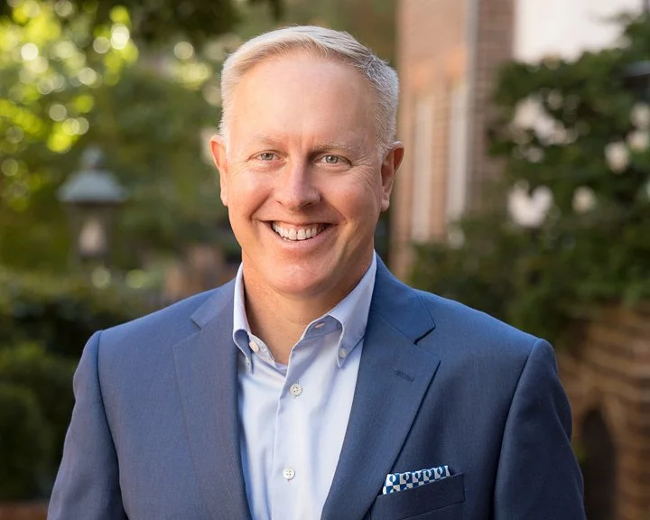 A middle-aged man with short gray hair smiling, wearing a blue suit and white dress shirt, standing outdoors with trees and a building in the background.