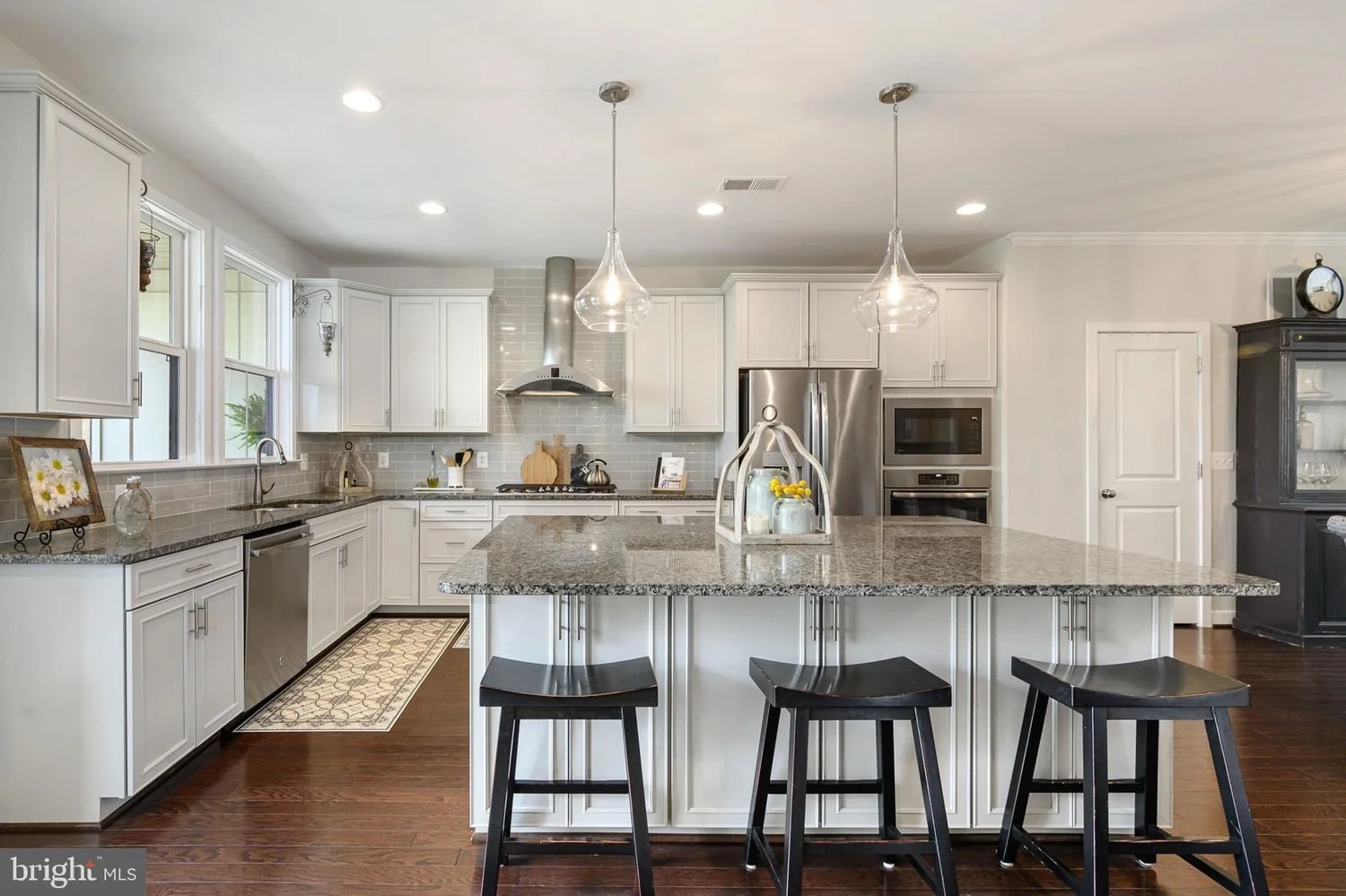 Modern kitchen with white cabinets, gray granite countertops, stainless steel appliances, dark wood flooring, and a central island with three black stools.