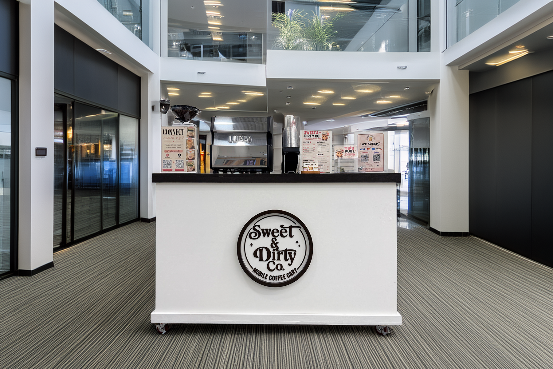 A mobile coffee cart named 'Sweet & Dirty Co.' located indoors in a modern building with glass and black accents, and a ceiling with yellow lighting.