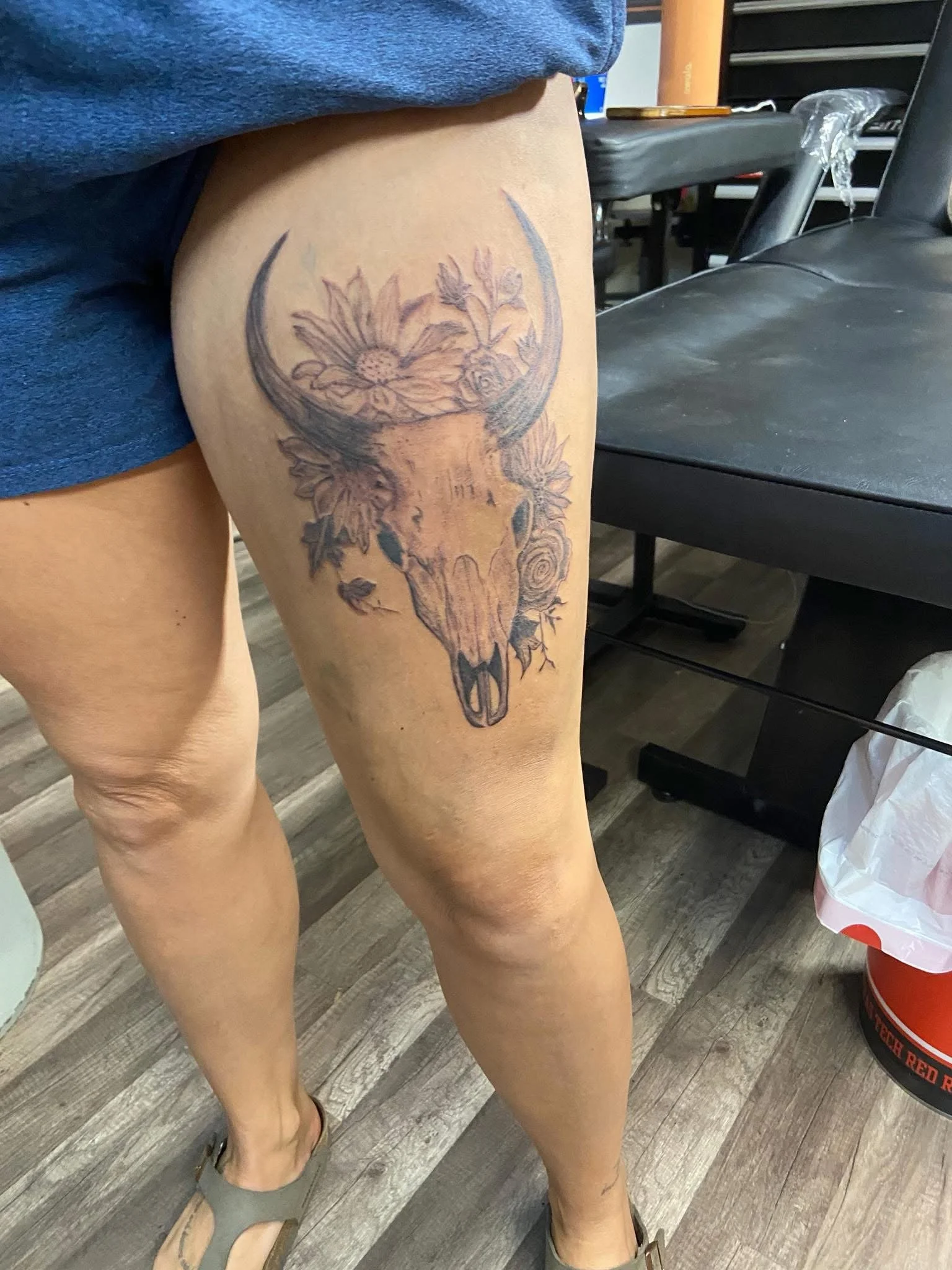 Tattoo of a deer's skull with flowers on the thigh of a person wearing shorts and sandals, standing on a wooden floor.