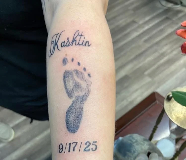 Tattoo of a baby footprint with the name 'Kashtin' above and the date '9/17/25' below.