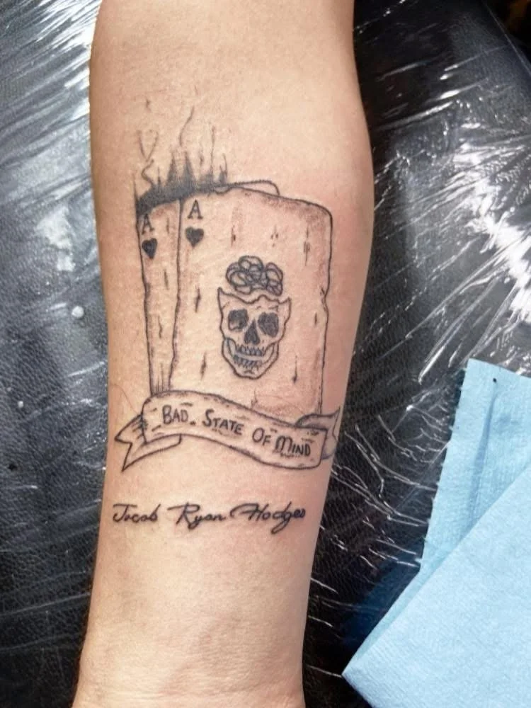 Tattoo of two playing cards showing aces, one partially burned with smoke, a skull with an intricate design, a banner with the text 'BAD STATE OF MIND,' and the name 'Jacob Ryan Hodge' written below.