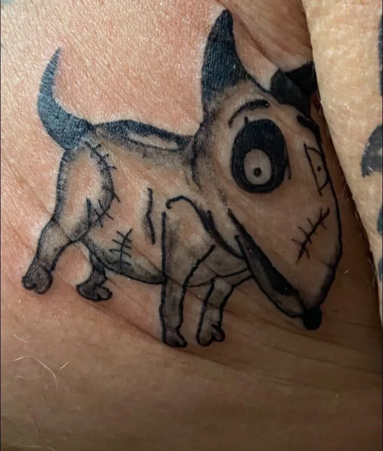 Tattoo of a cartoonish, dog-like creature with exaggerated features and big eyes on skin.