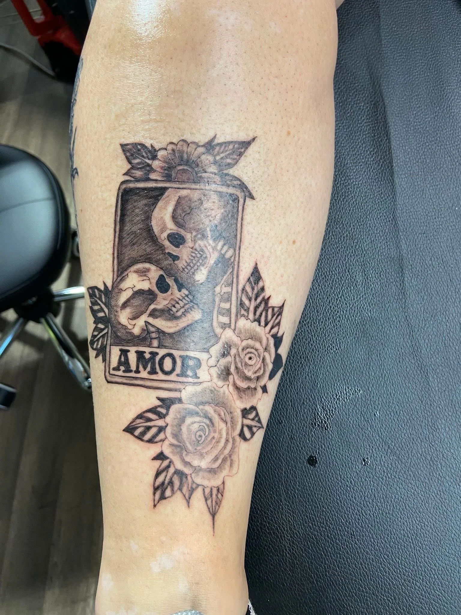 Tattoo on person's arm featuring a skull inside a rectangular frame, surrounded by roses and leaves, with the word 'AMOR' at the bottom.
