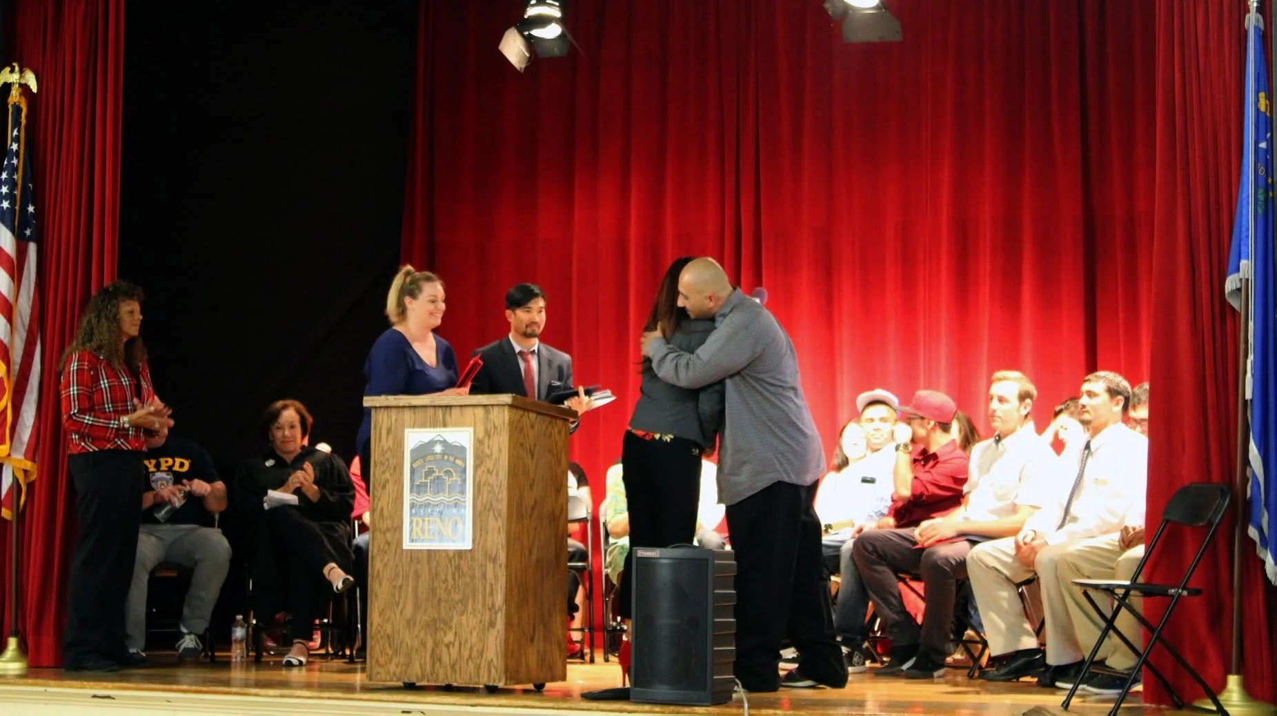 People on stage at a ceremony with red curtains, including someone hugging another person, a woman at a podium, and an audience seated in rows.