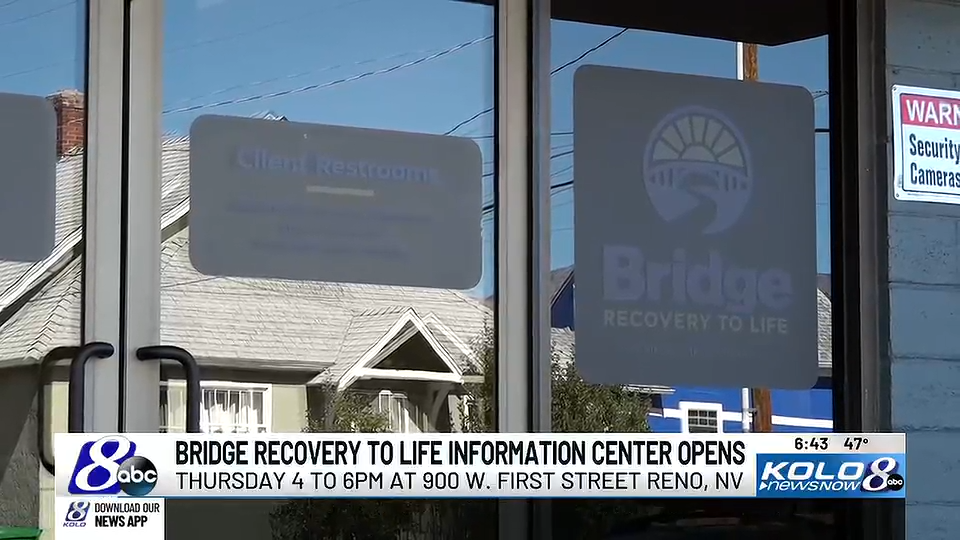 Close-up of glass door with signs showing the Bridge Recovery to Life center and a client restroom, with houses and power lines reflected in the glass.