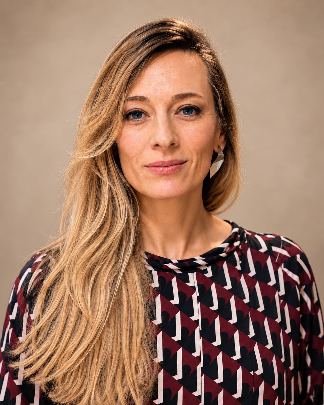 A woman with long blonde hair, blue eyes, and wearing a patterned blouse with geometric shapes, looks at the camera with a slight smile.