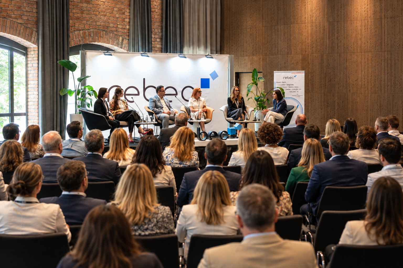 A panel discussion at a conference with six speakers on stage and an audience seated in front of them, in a modern room with brick walls and large windows.