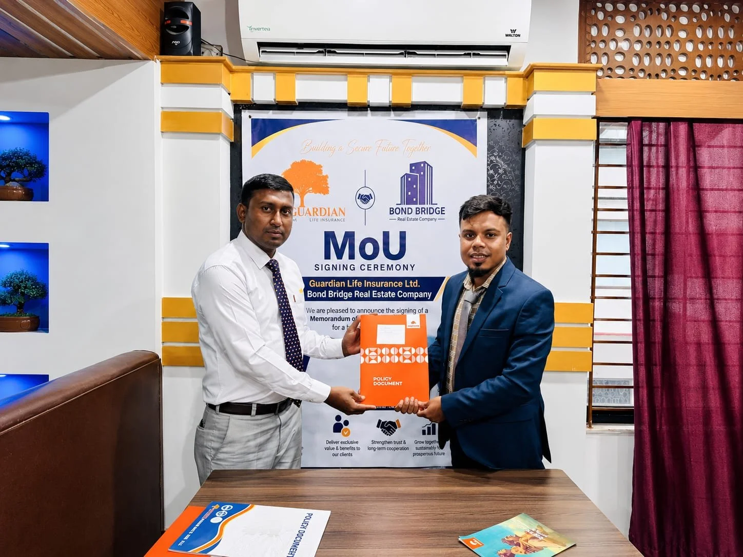 Two men in formal attire exchanging a policy document during a signing ceremony at an office with a banner in the background. The banner features logos and text related to Guardian Life Insurance Ltd. and Bond Bridge Real Estate Company.