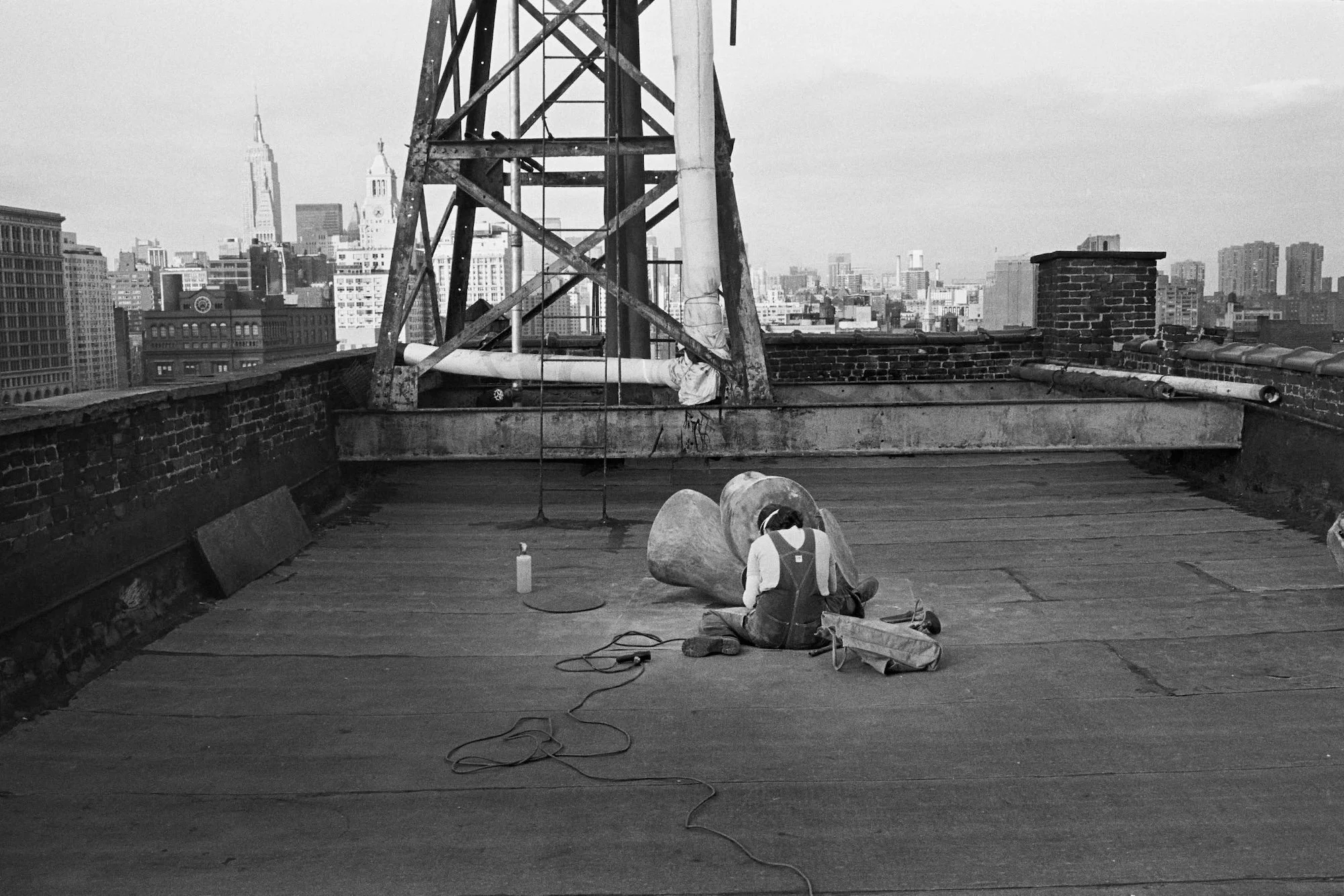 Person working on a rooftop construction or maintenance project, sitting on the floor with tools and equipment, with a city skyline in the background.