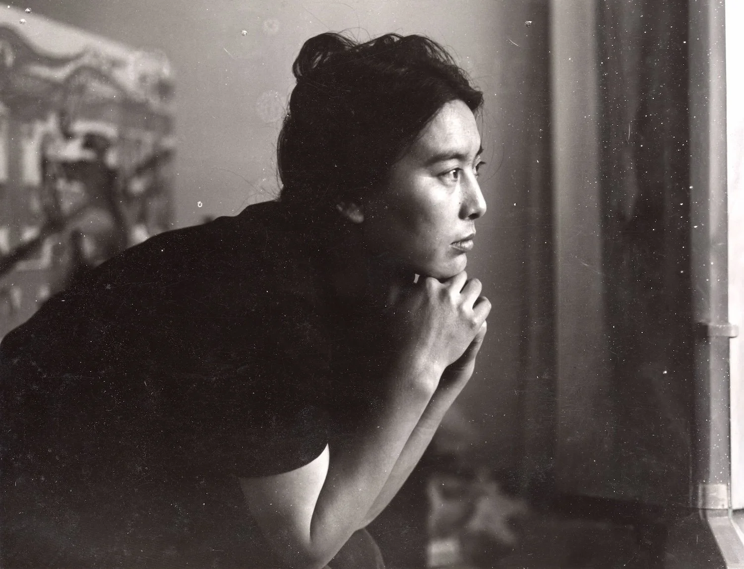 A black and white photo of a woman looking out a window with her hand under her chin, in a contemplative pose.