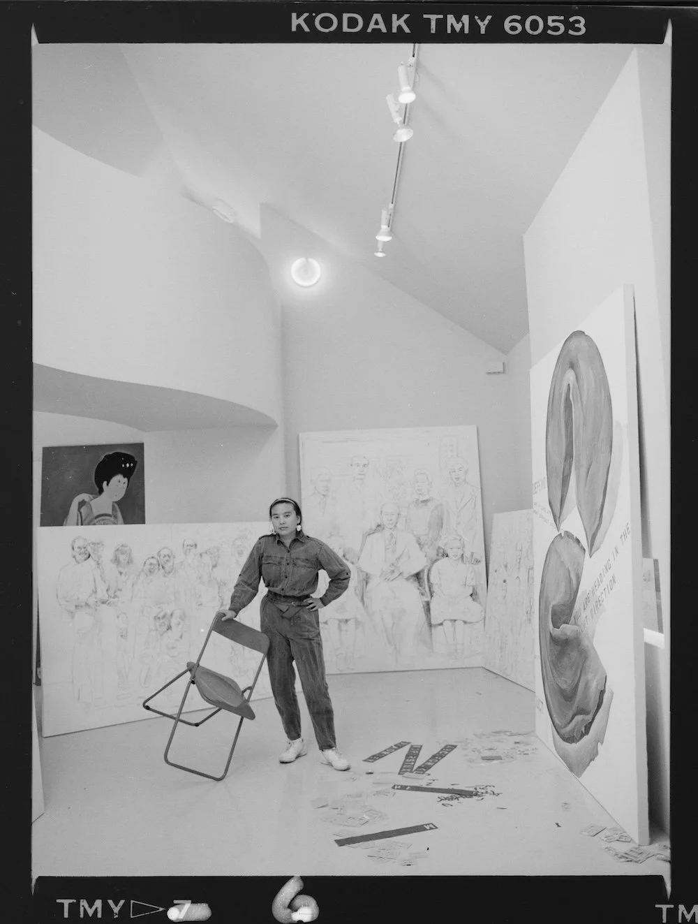 A woman standing in an art gallery with her hand on her hip, holding a folding chair. The gallery has various artworks, including sketches of people, a large leaf painting, and a portrait of a woman, displayed on the walls.