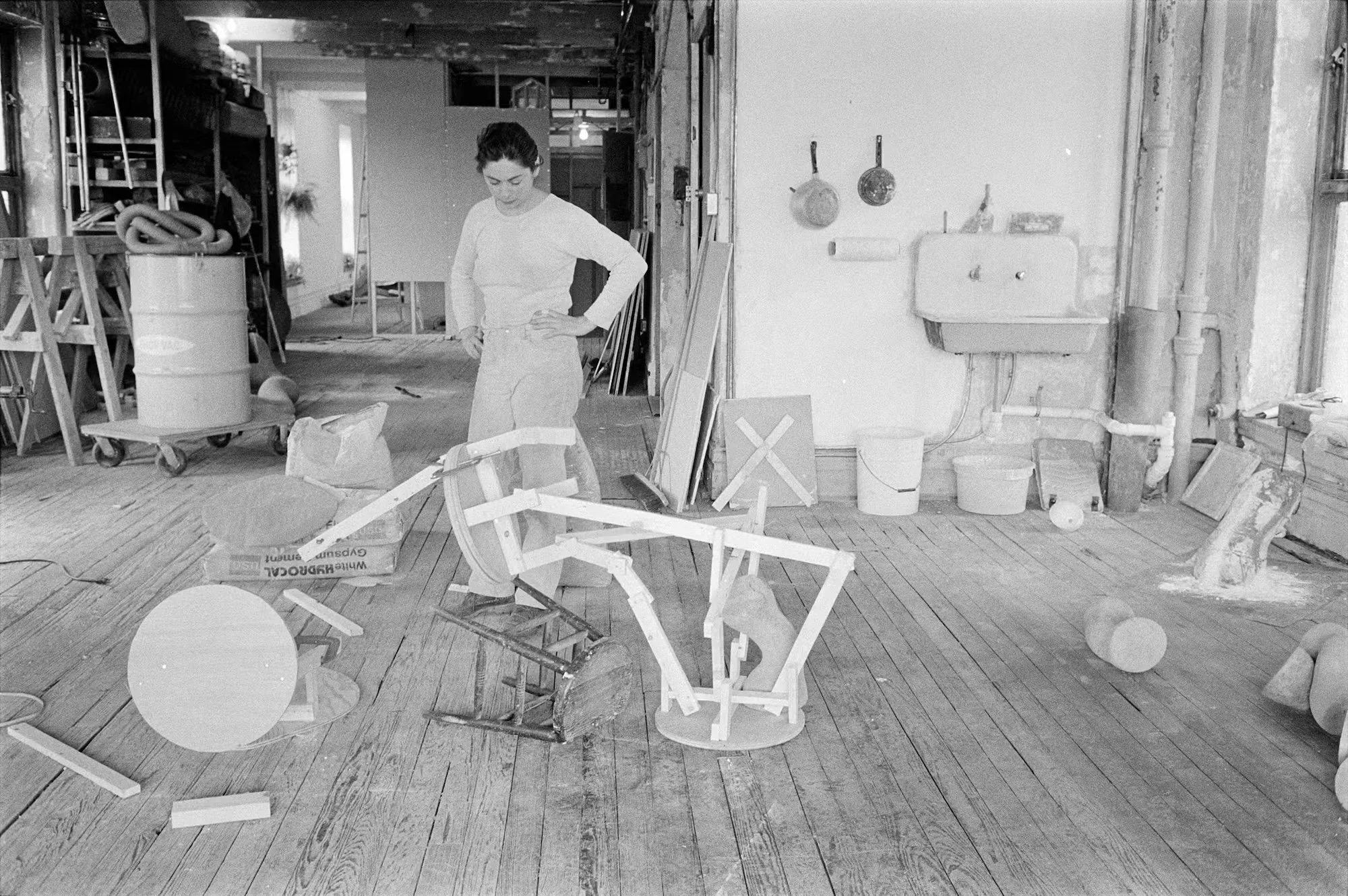 Woman in white shirt and pants working in a cluttered workshop or studio with various tools and objects, including a broken wooden chair, scattered on wooden floorboards.