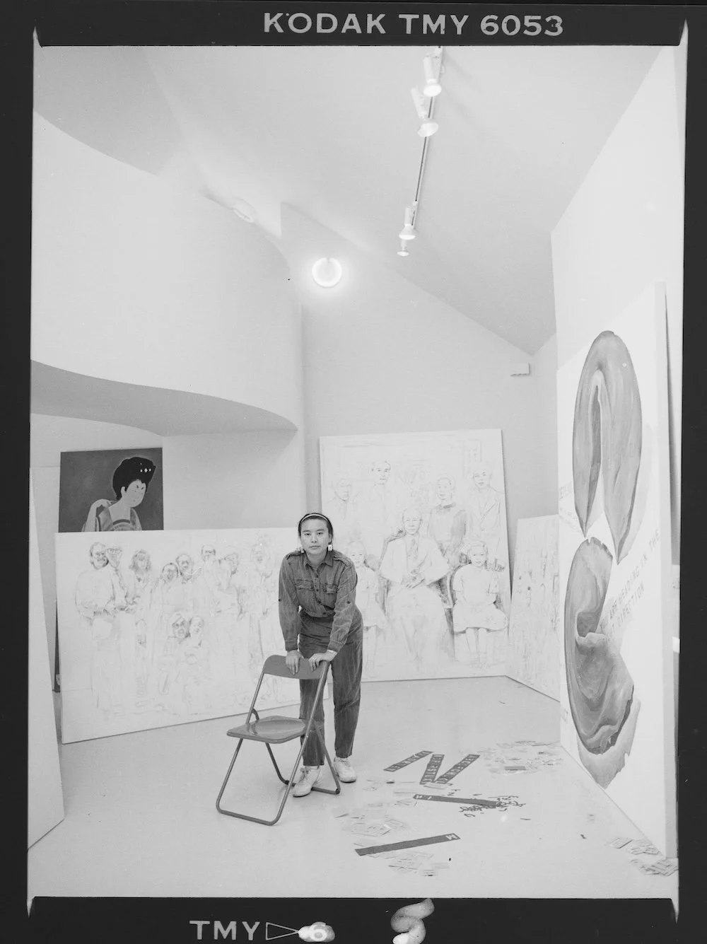 A person is standing and leaning on a chair in an art gallery. The background features various large paintings, including a portrait of a woman with styled hair on the left and a group portrait on panels behind the person. The gallery has high, curved ceilings with track lighting.