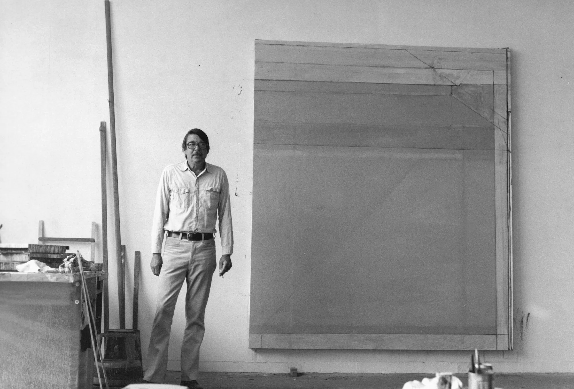 A man wearing glasses, a light-colored button-up shirt, and light-colored pants standing in an art studio or workshop, next to a tall, narrow wooden structure or sculpture. The background features plain walls, some art supplies, and a large wooden piece resting against the wall.