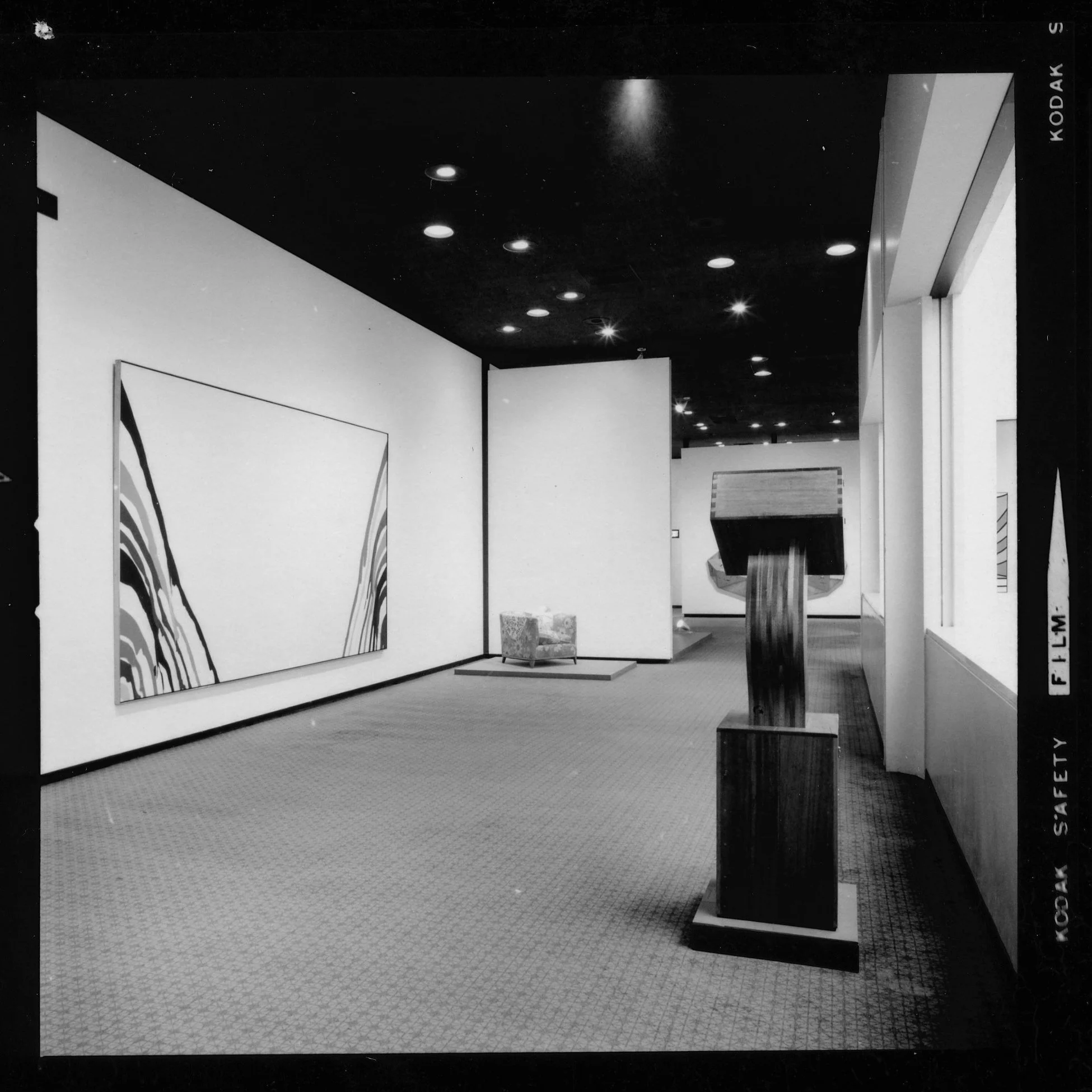 Black and white photograph of an art gallery with artworks on display, a display stand with a dark surface, and a patterned carpeted floor.
