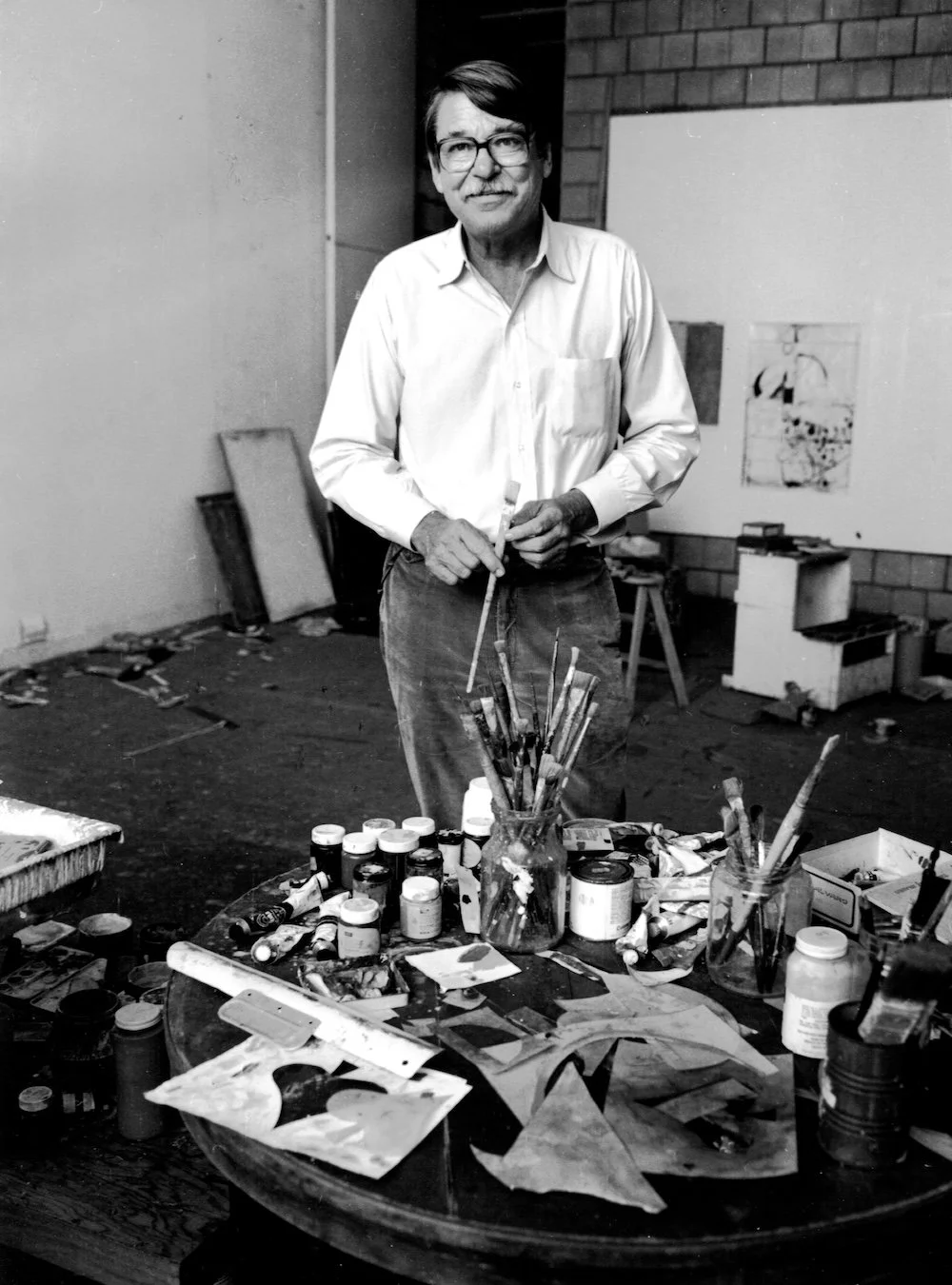 An older man with glasses in an art studio, standing behind a cluttered table with paint jars, brushes, and art supplies, holding a paintbrush.