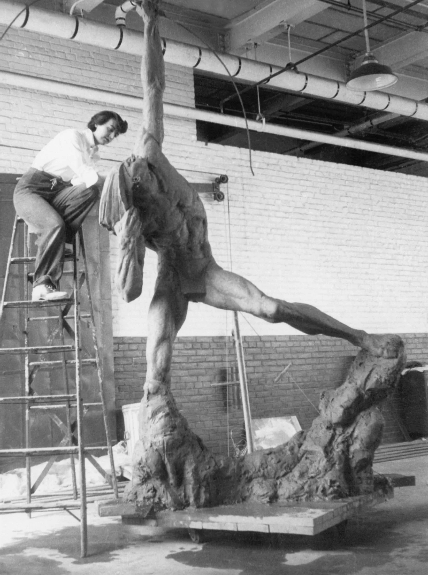 A woman working on a large sculpture of a lion in an indoor studio. The woman is sitting on a chair, and the sculpture is positioned on a platform with rocks and trees as part of the artistic design.