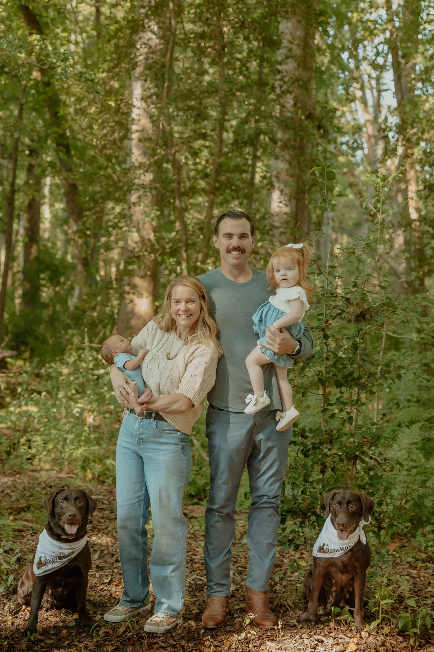 A family of four with two dogs standing in a wooded park, two adults with a woman holding a baby and a man holding a young girl, all smiling at the camera.