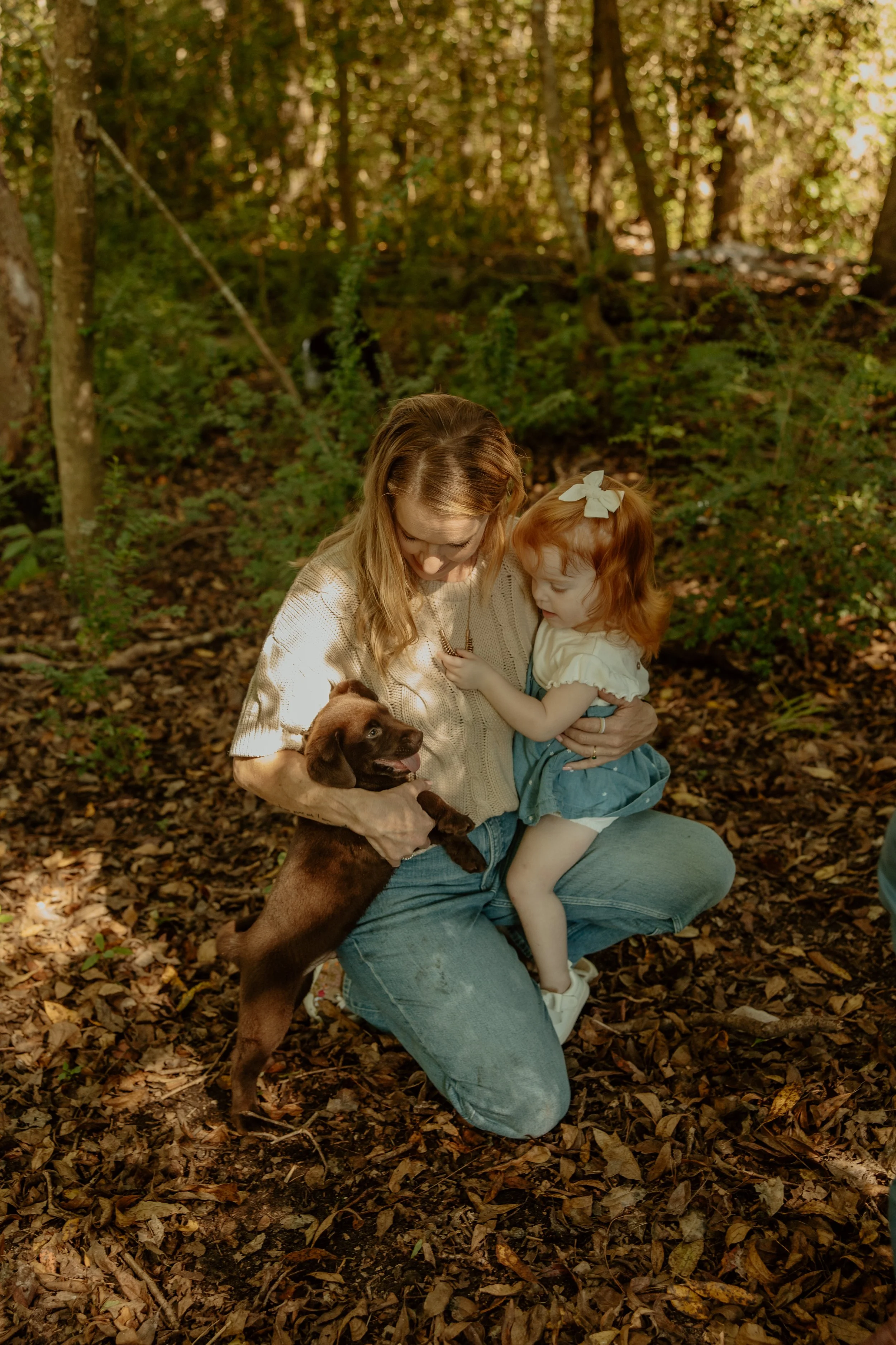 A woman with long blonde hair and a young girl with red hair in a bow are sitting outdoors in a wooded area. The woman is holding a brown puppy, and the girl is touching the puppy's face. The ground is covered with fallen leaves, and trees surround them.