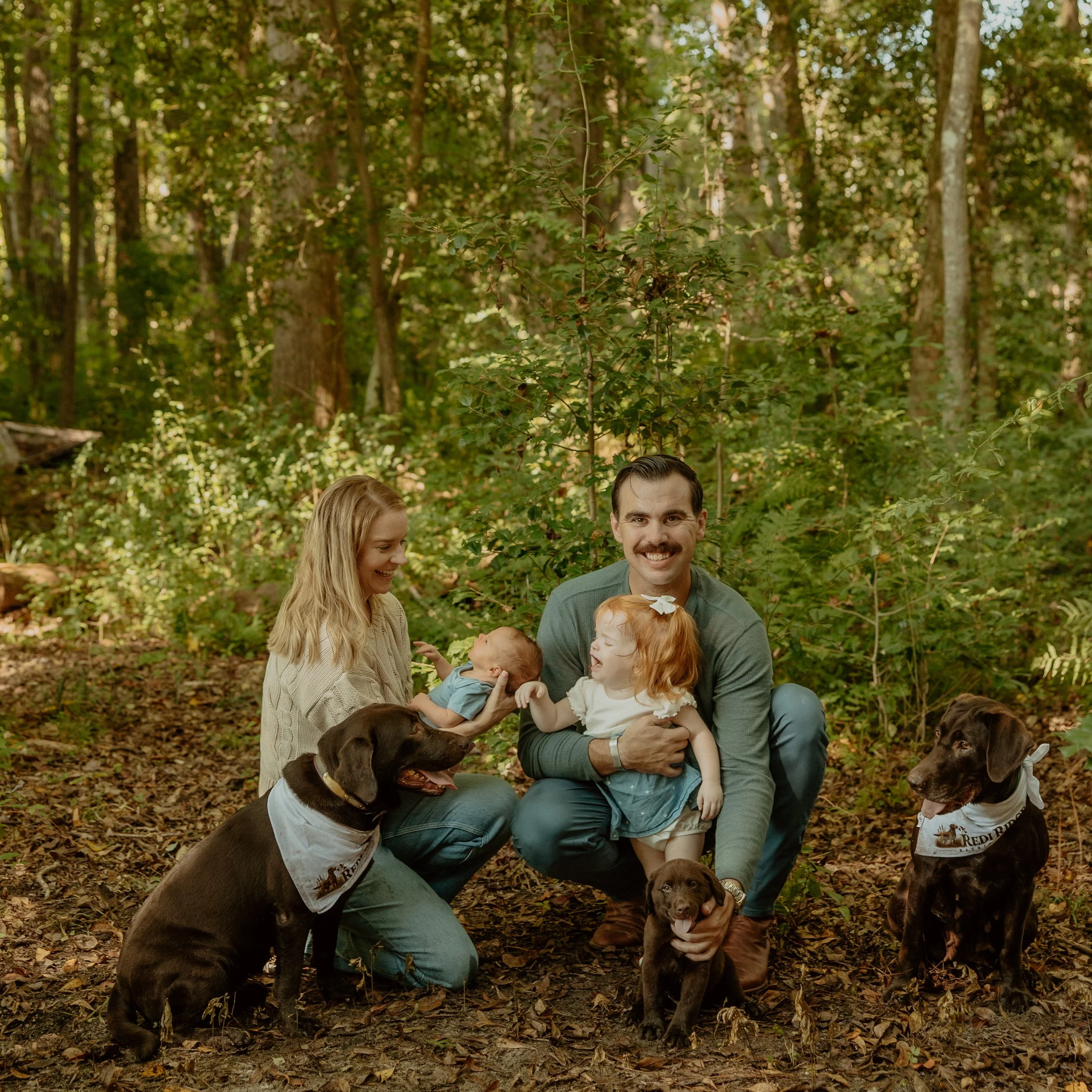 A family of four with two dogs outdoors in a wooded area. The woman has long blonde hair and is holding a small child. The man, with short dark hair, sits on the ground smiling, with a young girl with red hair and a white bow in her hair sitting on his lap. One dog is a brown Labrador with a white shirt, and the other is a black Labrador with a white shirt, sitting on the ground.
