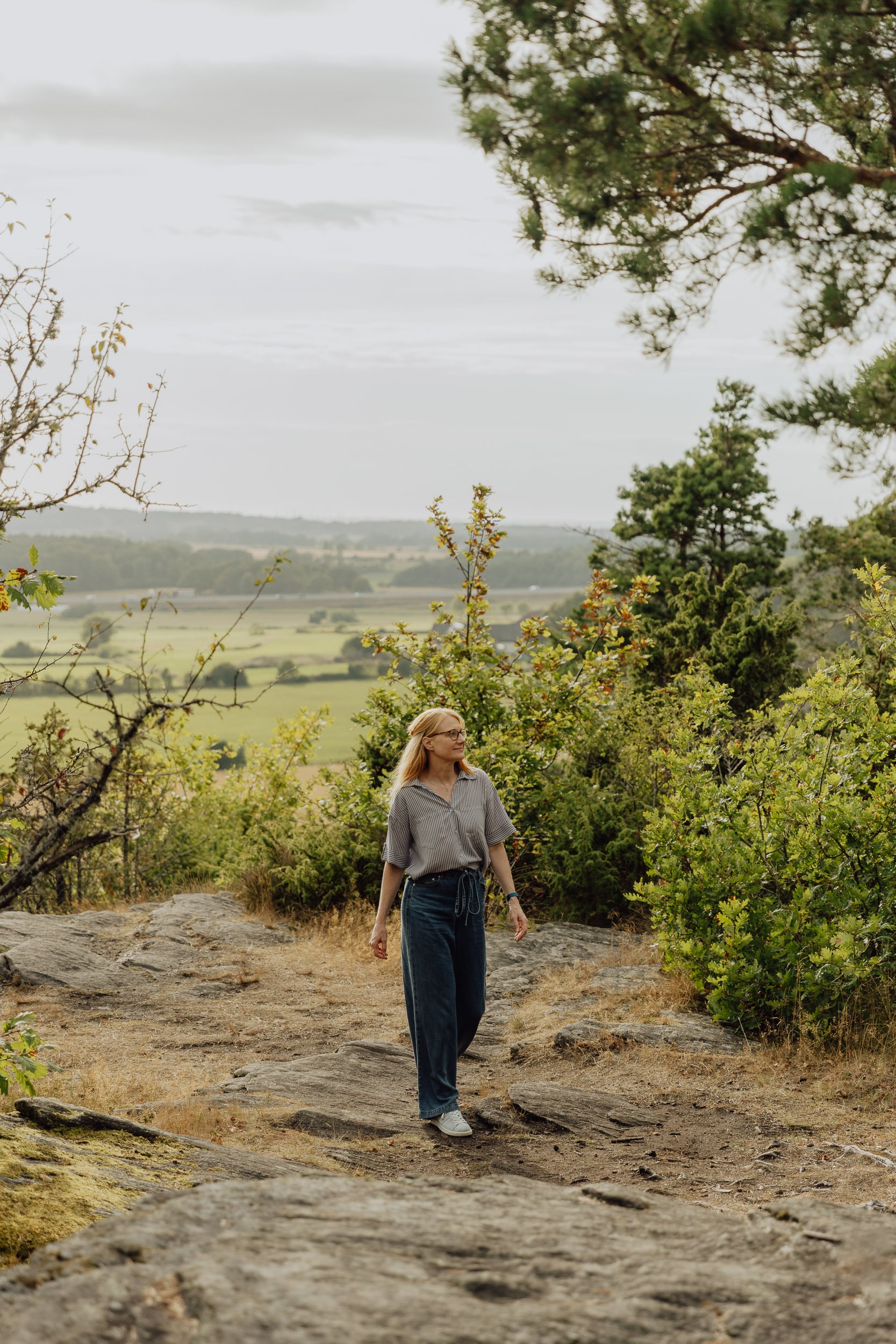 A woman with long blonde hair, glasses, wearing a striped shirt and jeans, walking outdoors on rocky terrain surrounded by greenery and trees, with a view of open fields and hills in the background on an overcast day.
