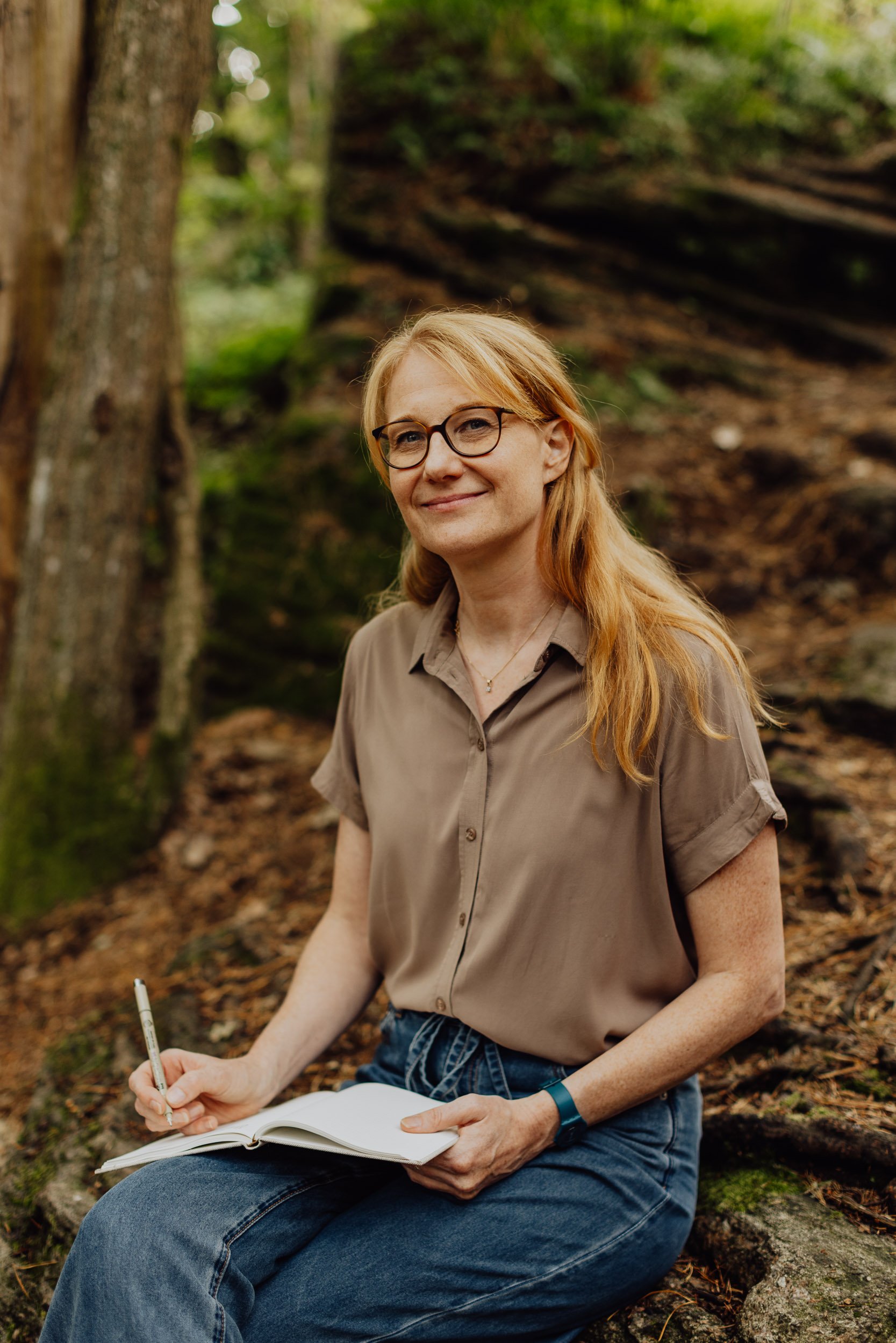 A woman with red hair, glasses, and a beige shirt sitting on a log in a forest, writing in a notebook.