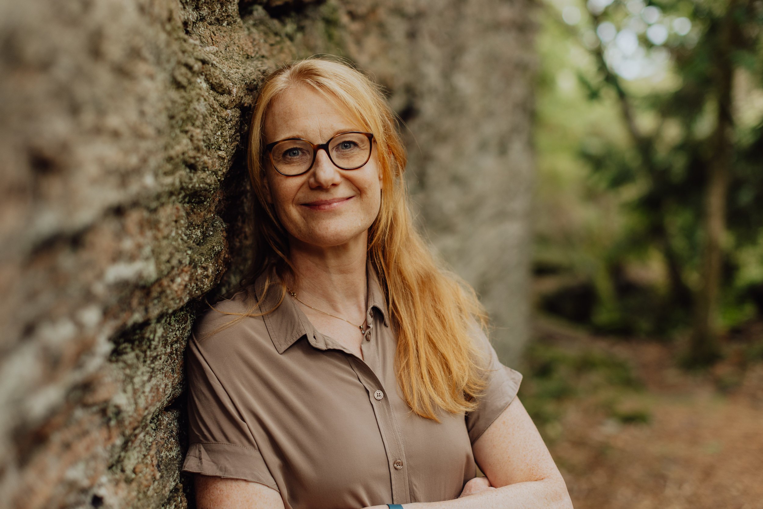 A woman with red hair and glasses smiling and leaning against a mossy brick wall outdoors with green trees in the background.