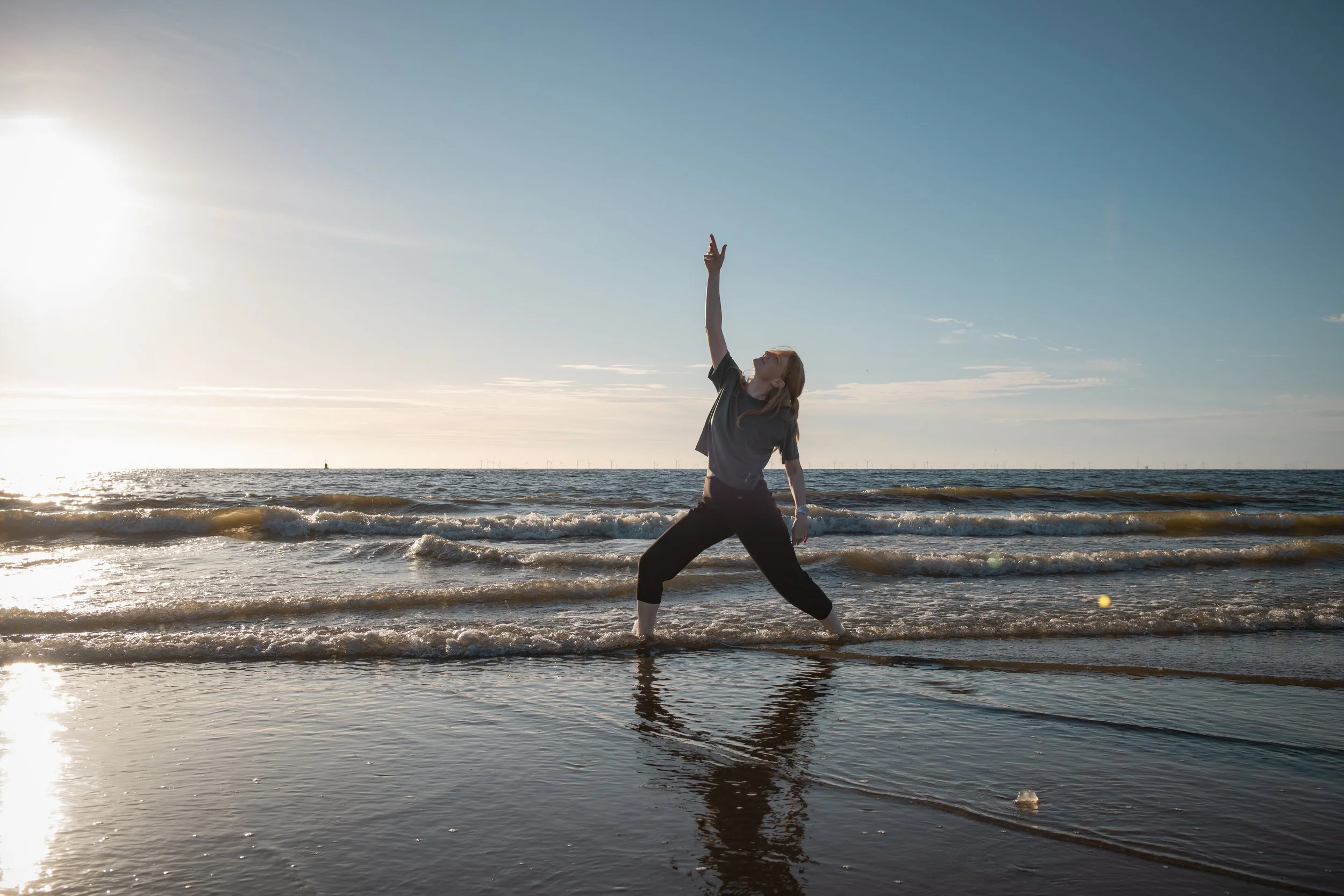 Woman practicing yoga on the beach during sunset, standing in a wide stance with one arm reaching up.