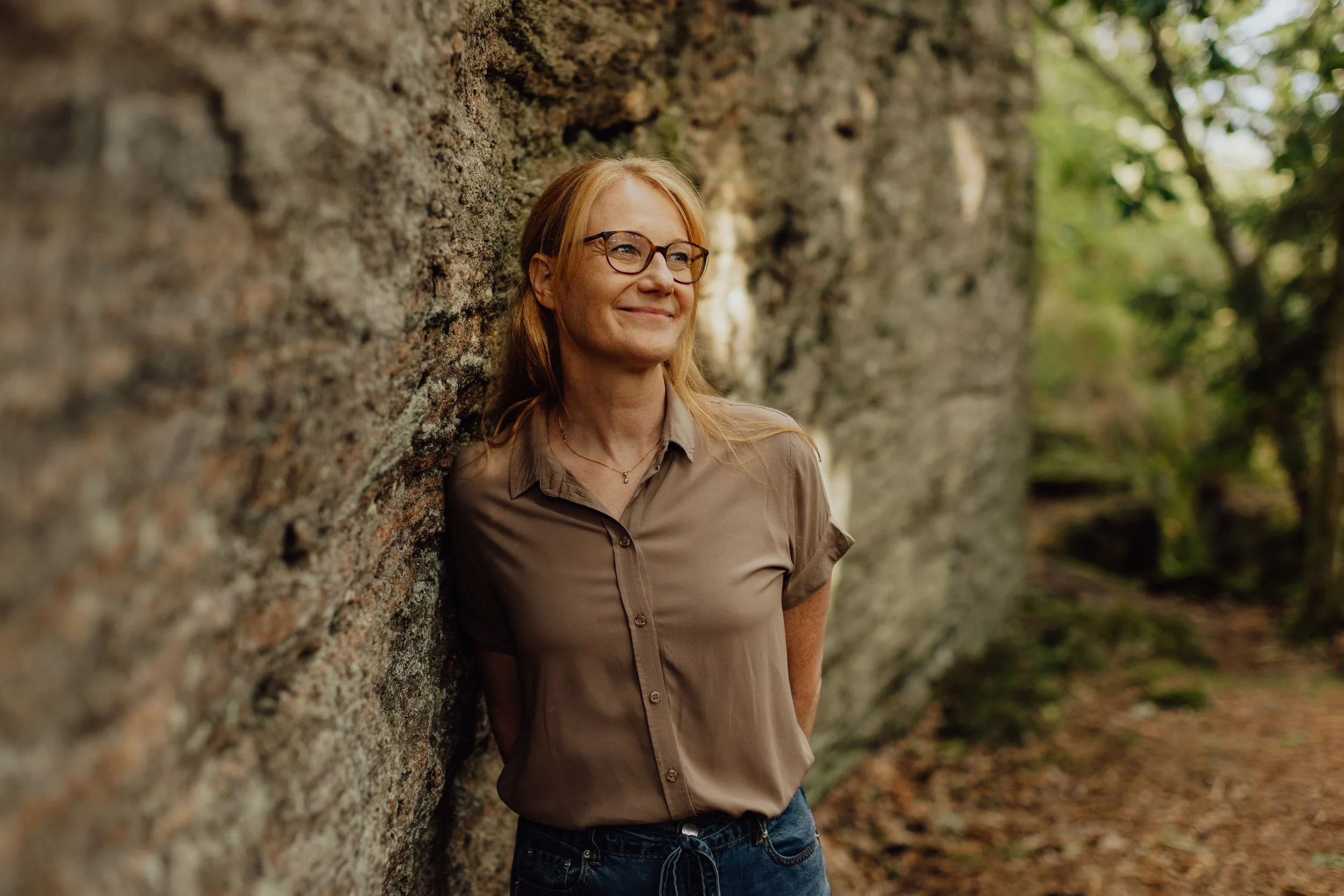 A woman with red hair and glasses standing outdoors near a stone wall, smiling and looking to the side in a natural, green setting.