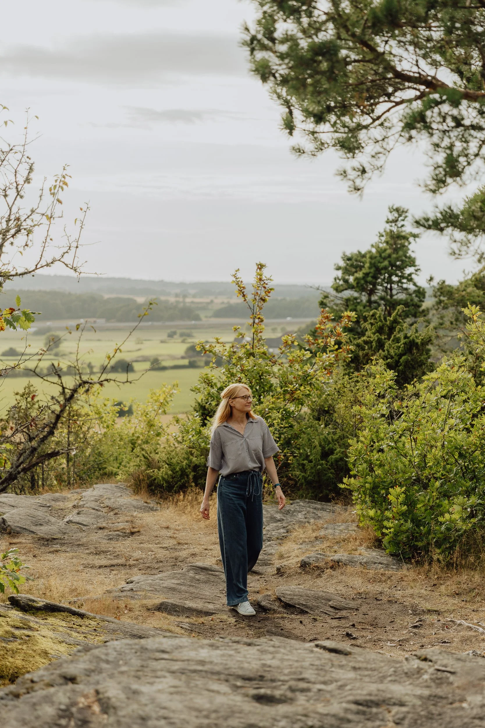 A woman with blonde hair, glasses, wearing a striped shirt and high-waisted jeans, walking on a rocky path in a lush green outdoor setting. Trees and rolling farmland are visible in the background under an overcast sky.