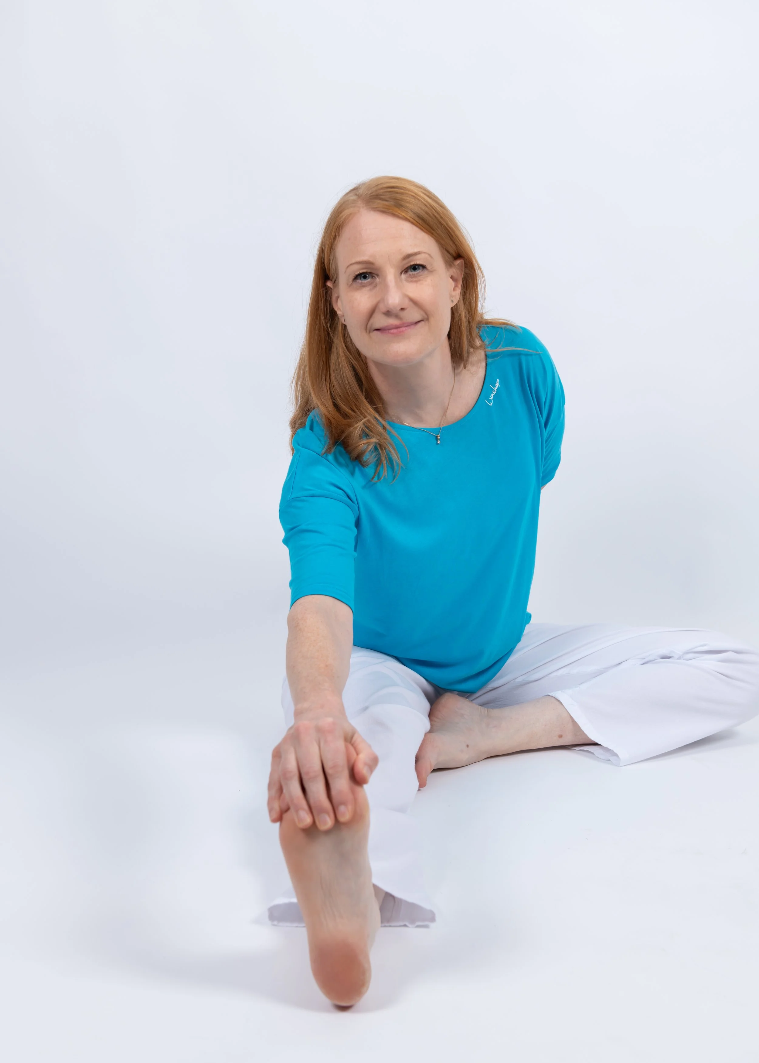 A woman with red hair wearing a blue top and white pants, sitting cross-legged on the floor, reaching forward with her right hand and holding hands with another person.