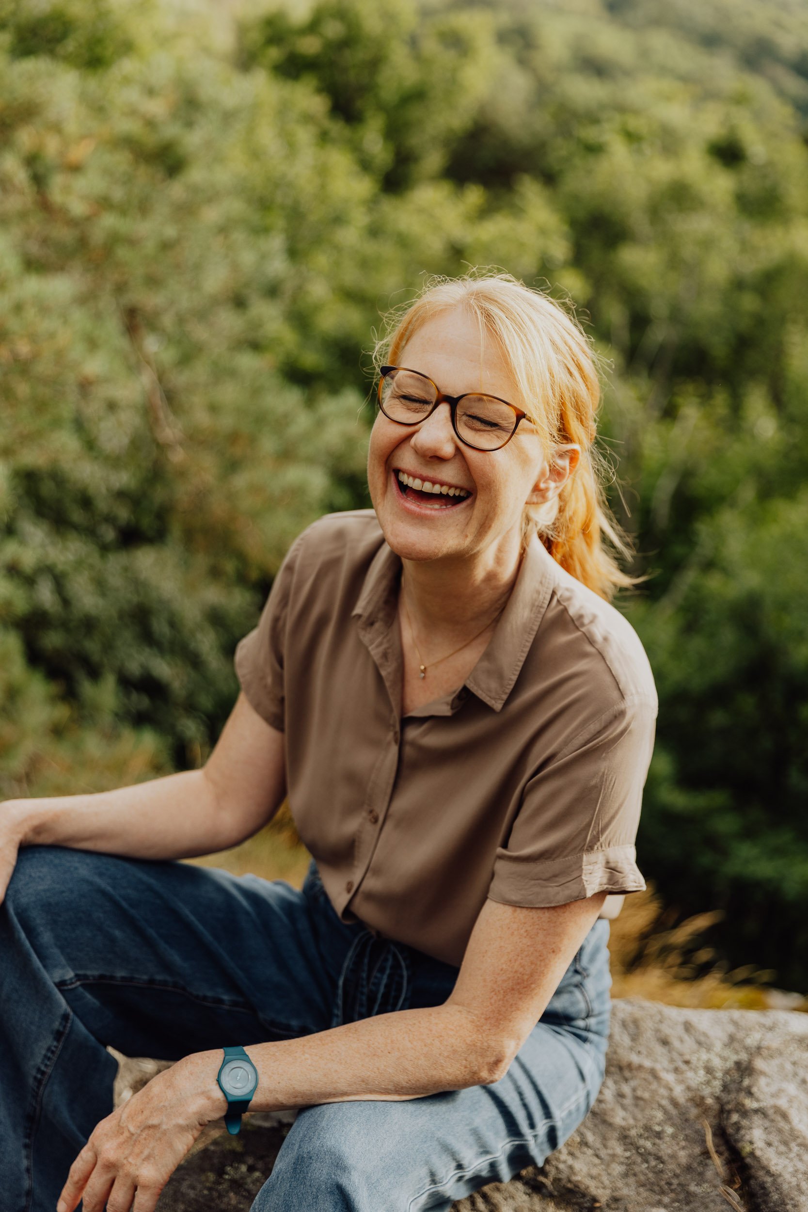 A smiling woman with glasses and red hair, wearing a brown shirt and blue jeans, sitting outdoors on a rock with green foliage in the background.