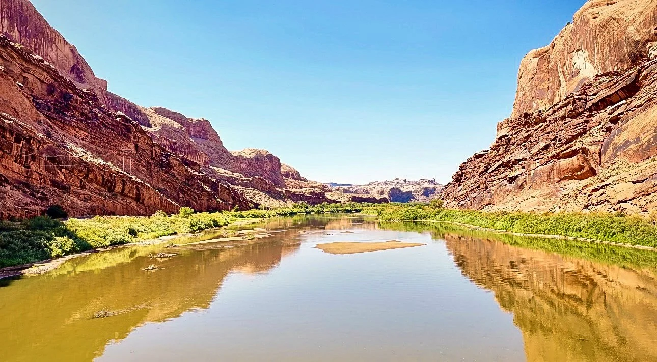 A river flowing through a canyon with red rock walls on both sides and green vegetation along the banks, under a clear blue sky.