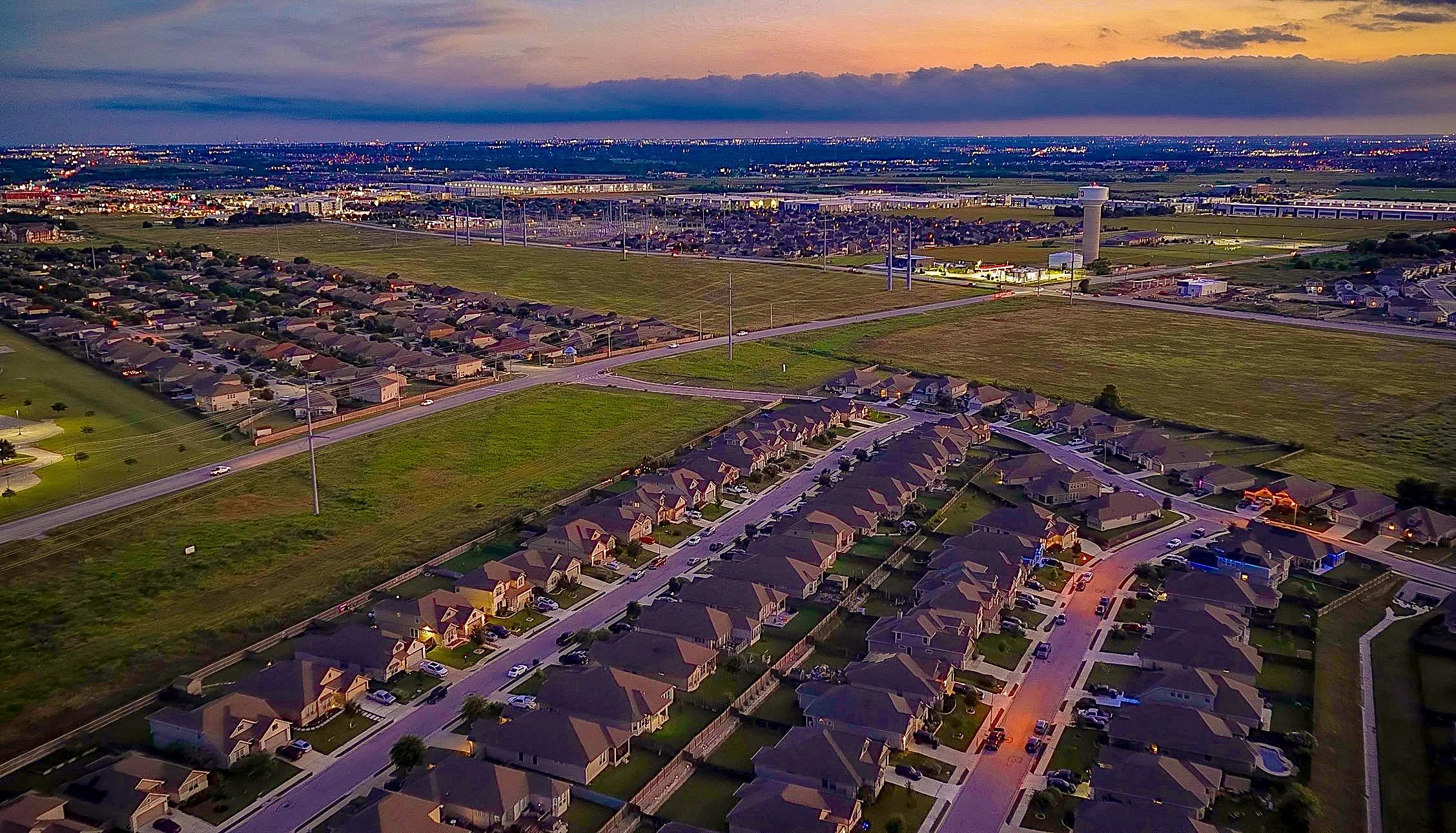 An aerial view of a suburban neighborhood at dusk, showing houses with lit windows, streets, and a flat open field with wind turbines. In the background, a cityscape and a dark horizon under a colorful sky.