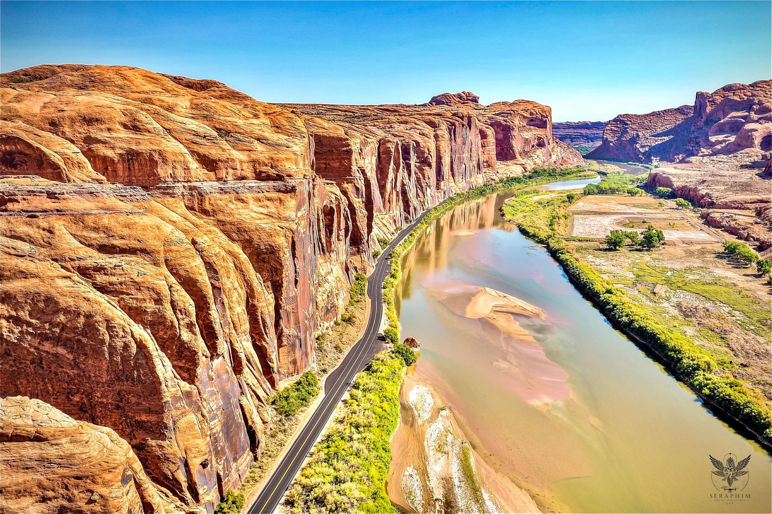 Colorful canyon landscape with steep orange and red rock formations on the left, a winding river in the middle, and green vegetation and fields on the right under a clear blue sky.