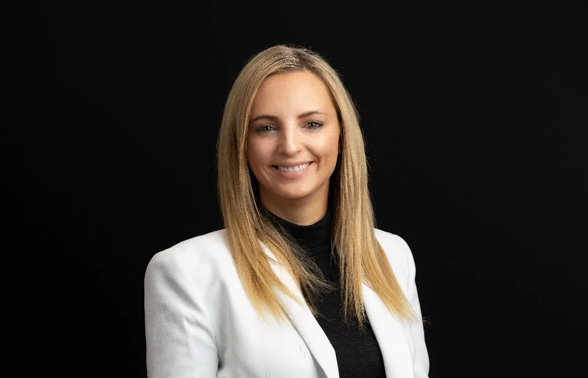 Portrait of a woman with blonde hair wearing a white blazer and black top against a dark background.