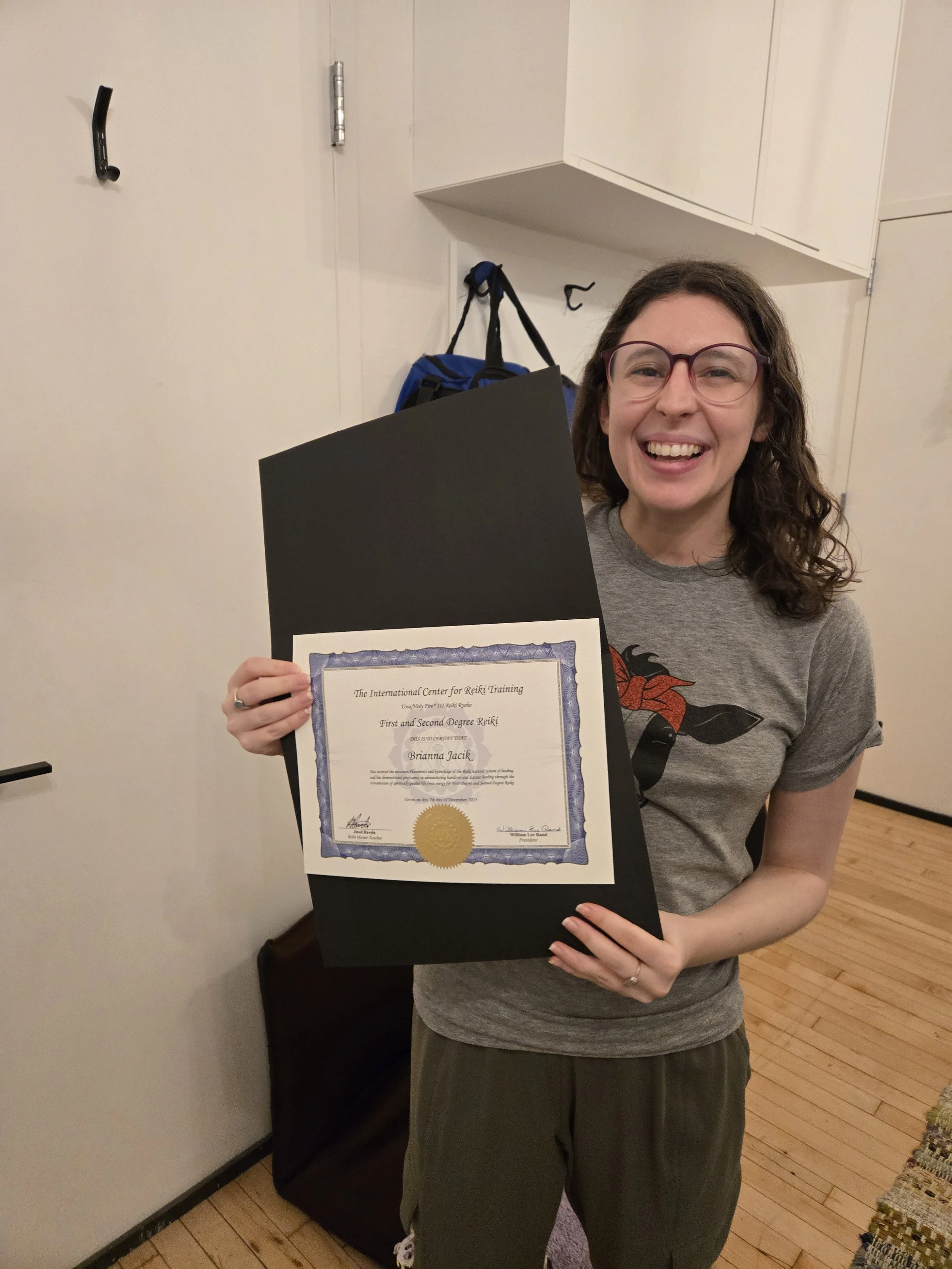 A woman with glasses is smiling and holding a certificate of achievement for First and Second Degree Reiki. She is standing indoors near a white door and a white cabinet, with a blue bag hanging on a hook behind her.