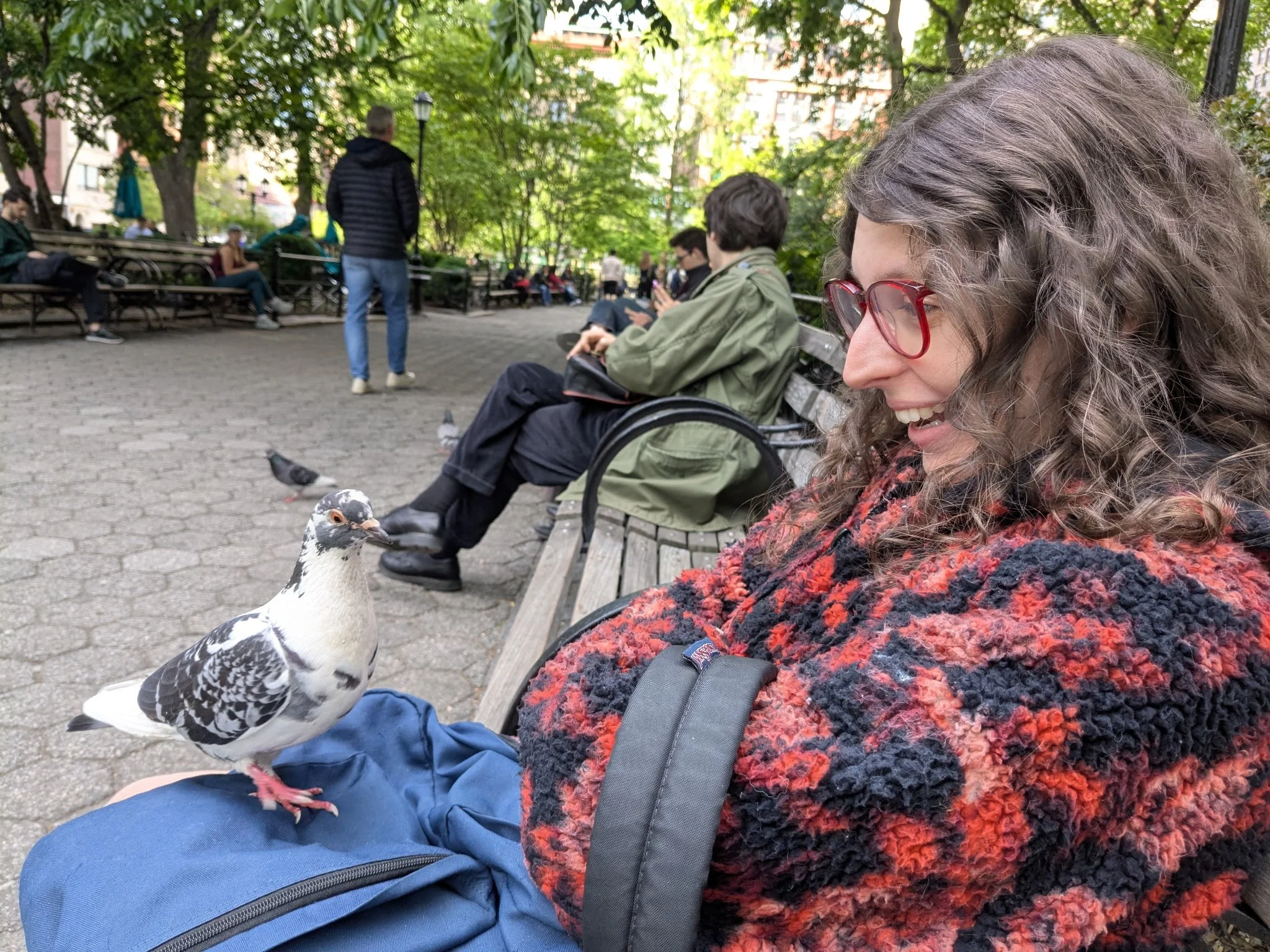 A woman with curly hair, red glasses, and a colorful sweater sitting on a park bench, smiling at a pigeon on her leg, with people sitting and walking in the background under trees in a city park.