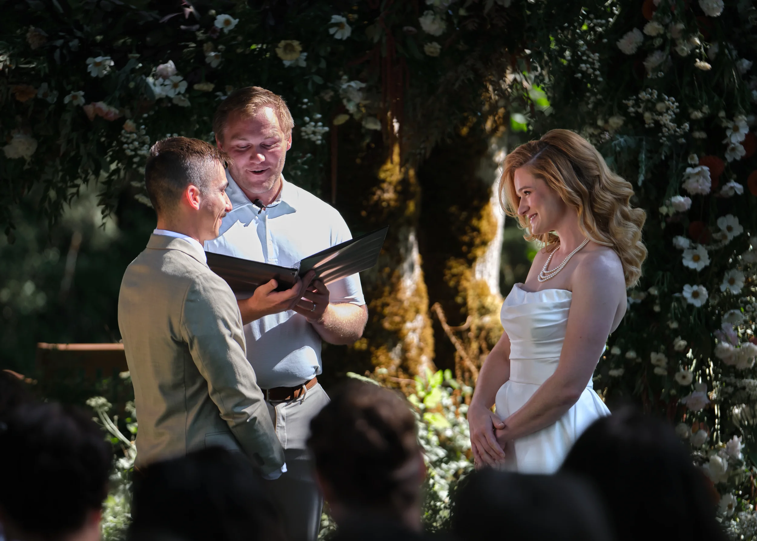 A wedding ceremony with a couple facing each other, exchanging vows, with an officiant reading from a book. The bride is wearing a strapless white wedding dress and pearl necklace, and the groom is wearing a beige suit. The background is decorated wi
