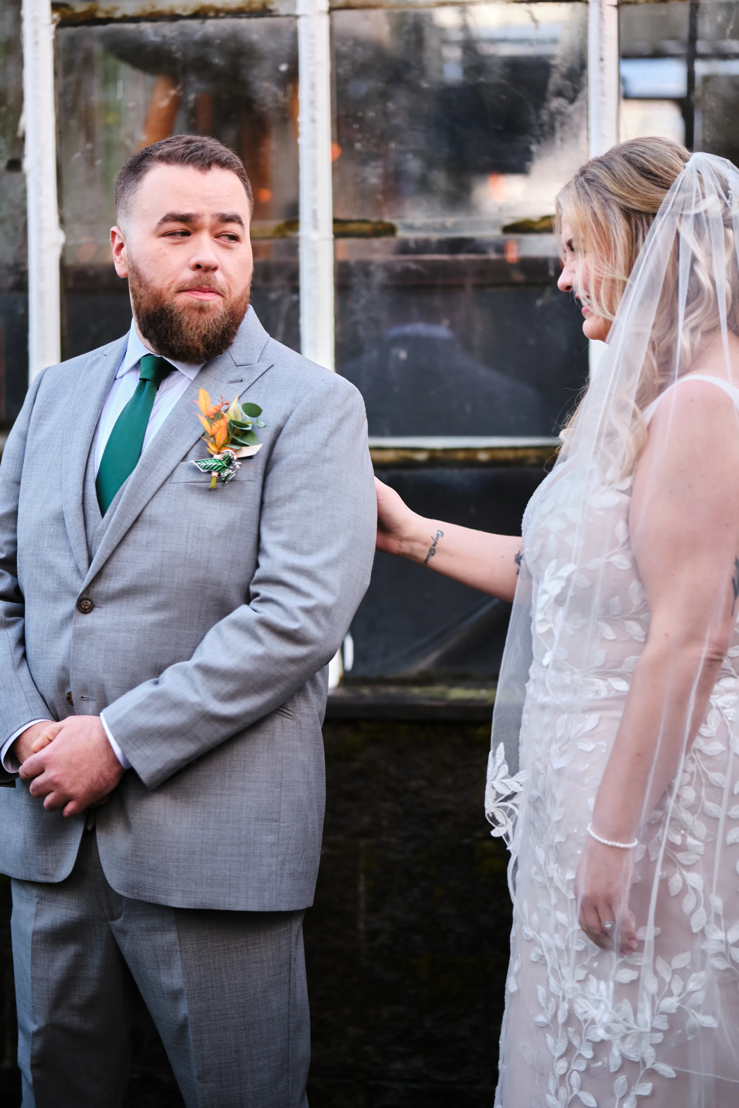 A groom in a gray suit and green tie with a boutonniere stands facing a bride, who wears a white lace wedding dress with lace floral patterns and a veil. The bride is gently touching the groom's shoulder and smiling softly, while the groom looks to t