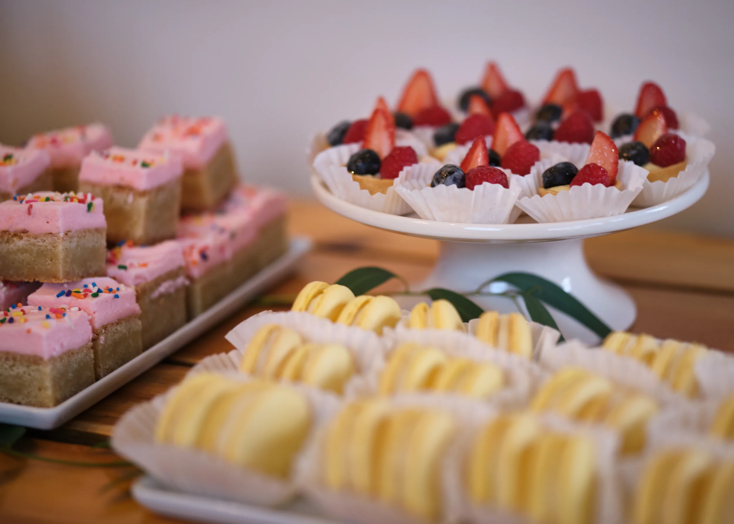 Assorted small desserts on white plates, including fruit-topped tartlets with strawberries, blueberries, and raspberries, and pink frosted cakes with rainbow sprinkles, arranged on a wooden table with green leaves for decoration.