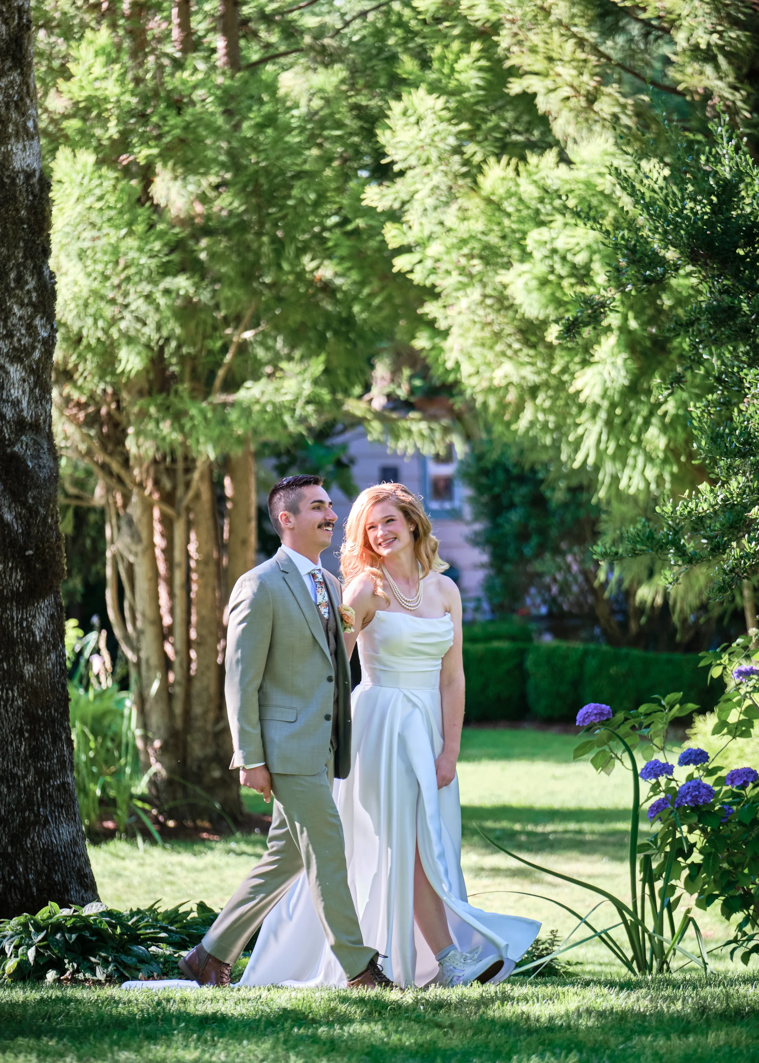 A smiling couple walking outdoors in a lush garden with trees and flowers, dressed in formal attire, suggesting a special occasion or celebration.