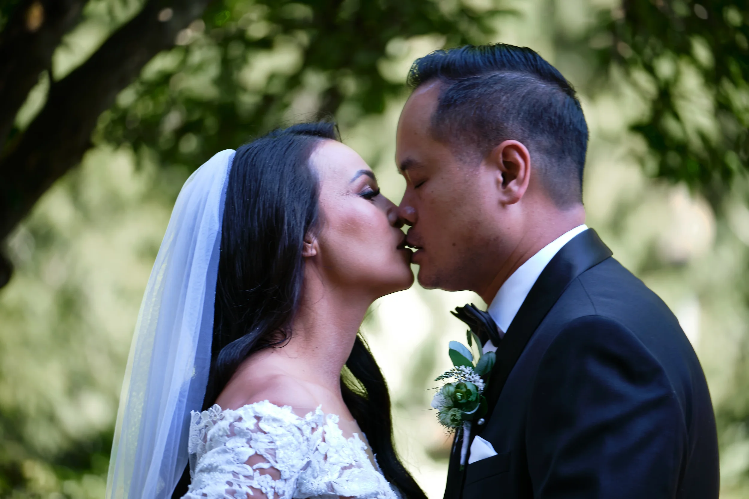 A bride and groom are touching noses outdoors during a wedding ceremony, with green trees in the background.
