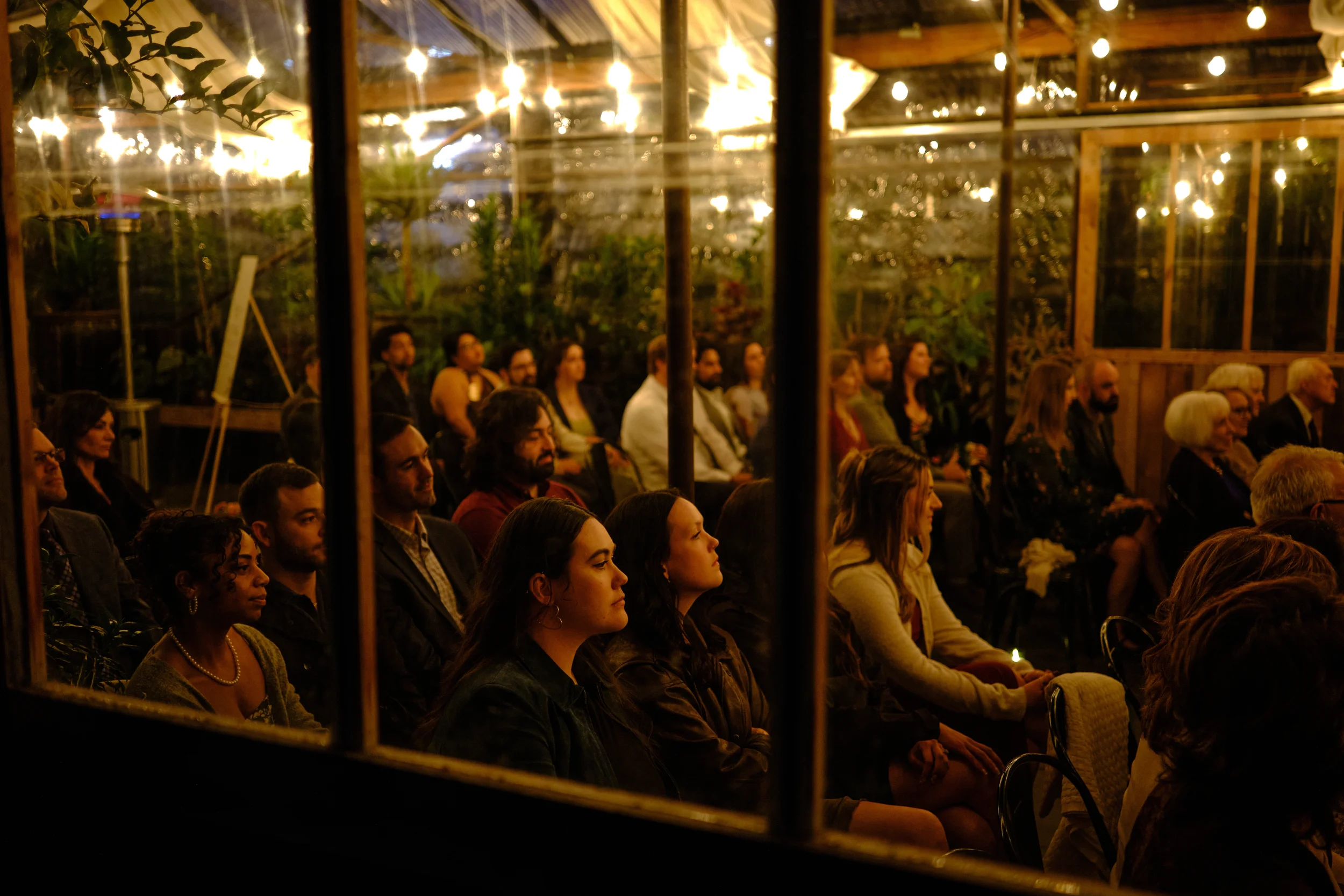 People seated indoors during an evening event, viewed through a glass window with visible reflections and warm lights overhead.