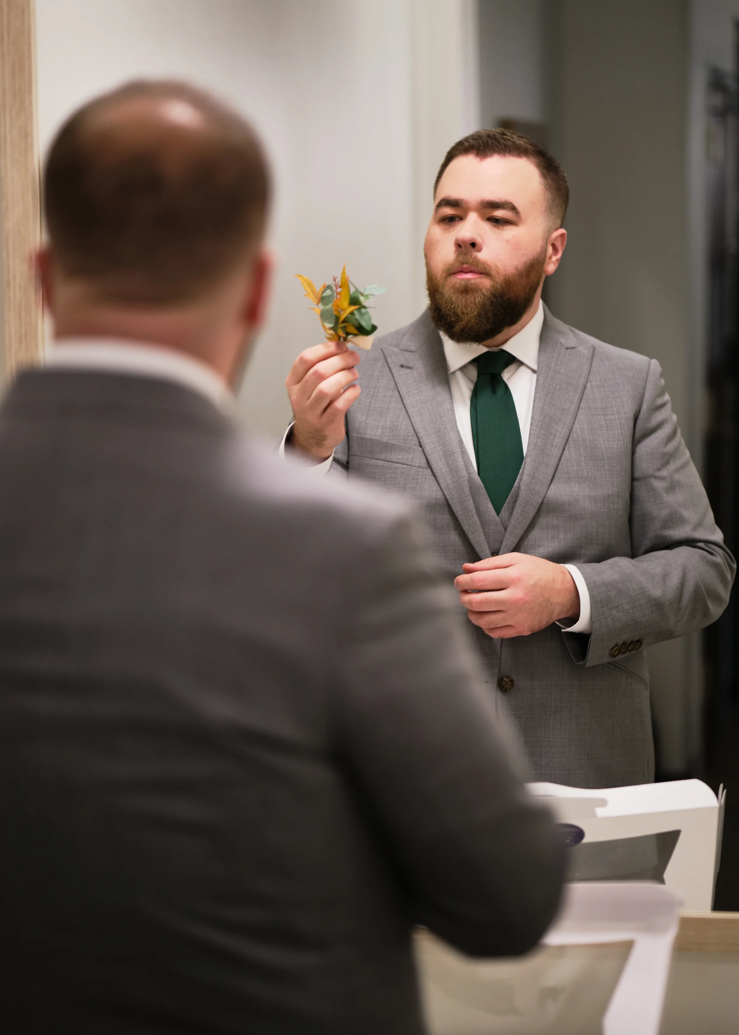 A man in a light gray suit with a dark green tie holding a boutonniere while looking at another man in a dark suit.